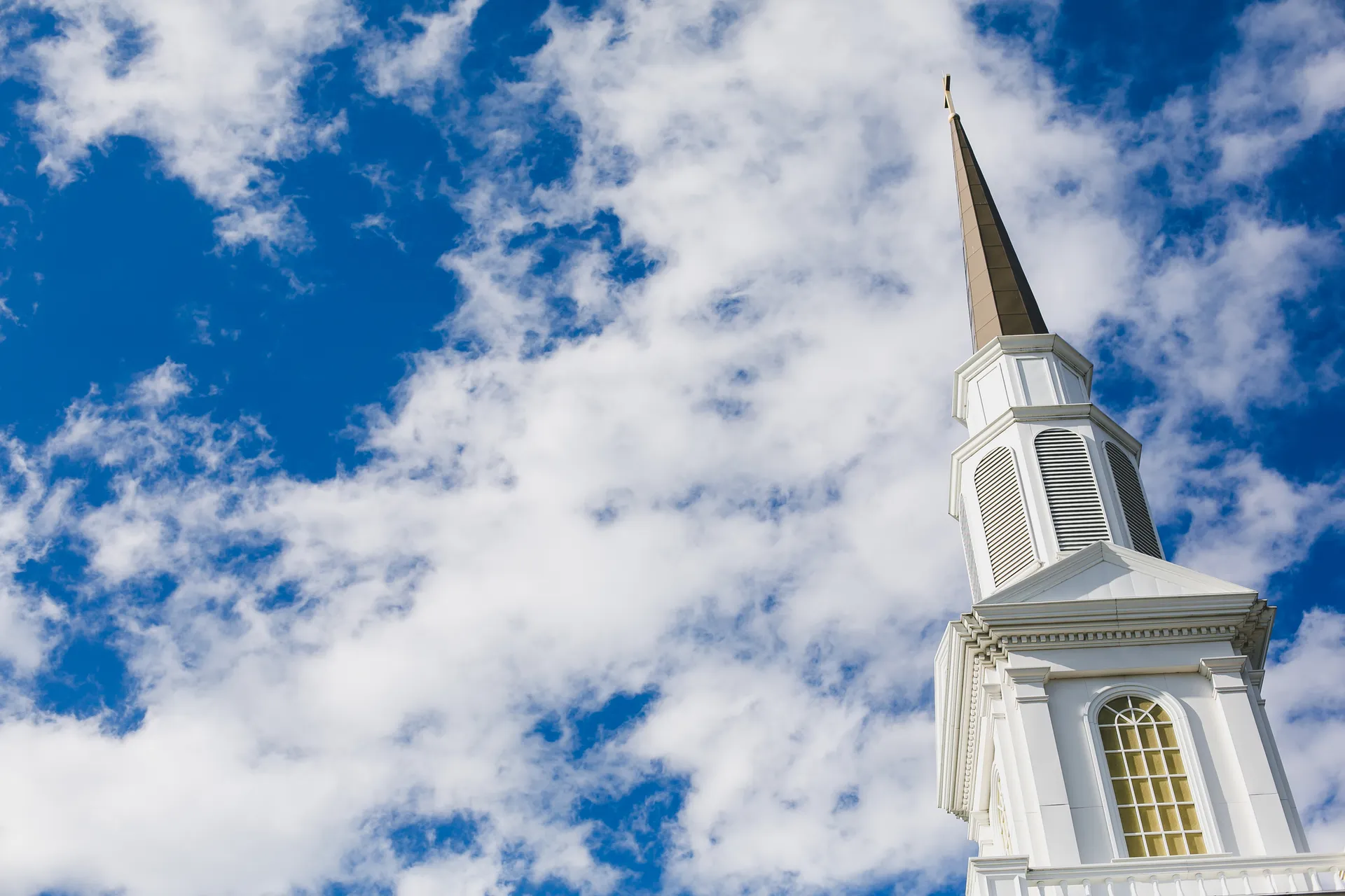 A white church steeple with a cross reaching into a blue sky with clouds